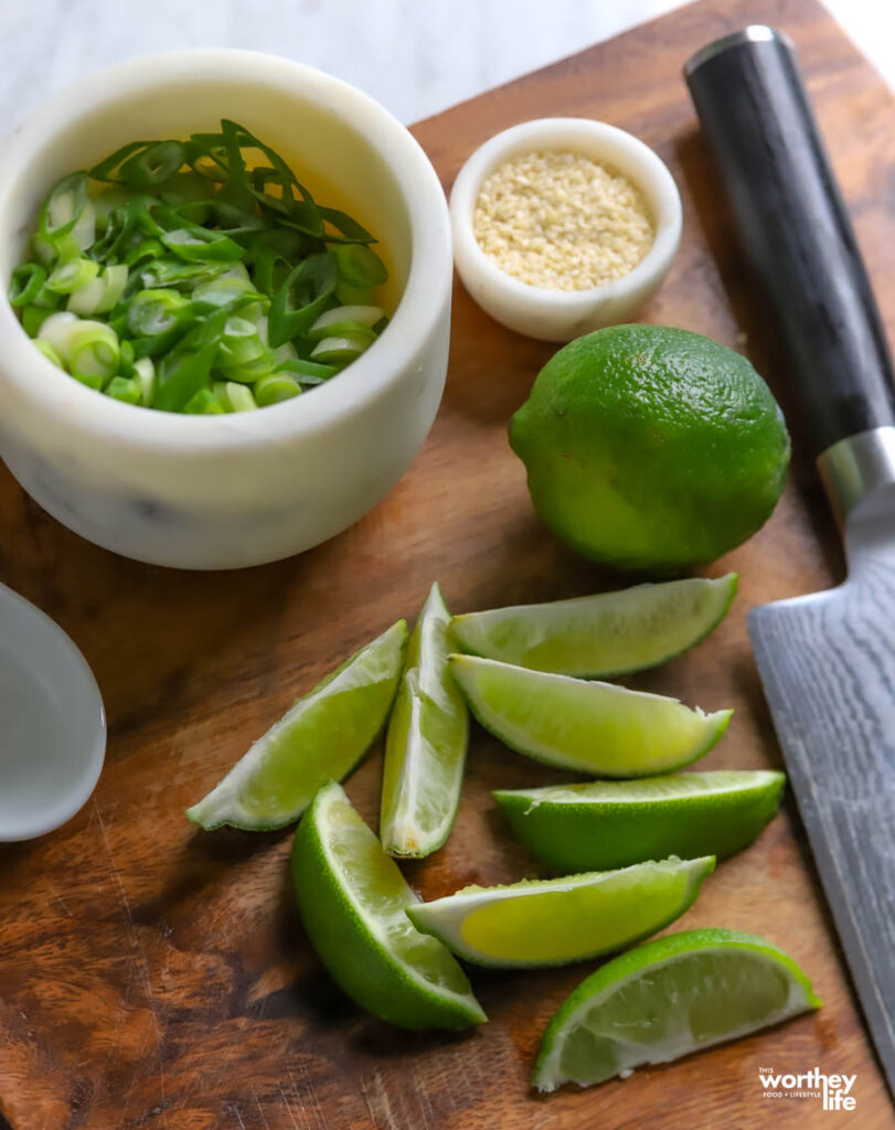 a cutting board loaded with cut limes, green onion, sesame seeds and a knife.