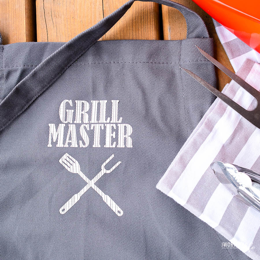 Grey DIY apron with white "grill master" message on front laying on table by grill utensils