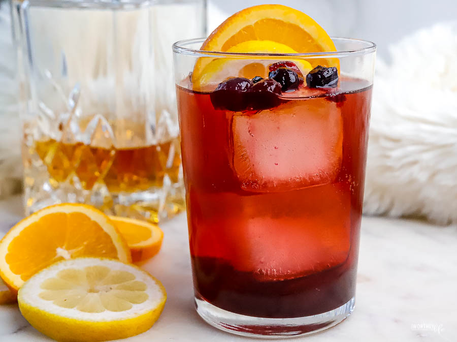 a glass filled with a reddish cocktail mixture with ice, berries, and slices of fruit. 