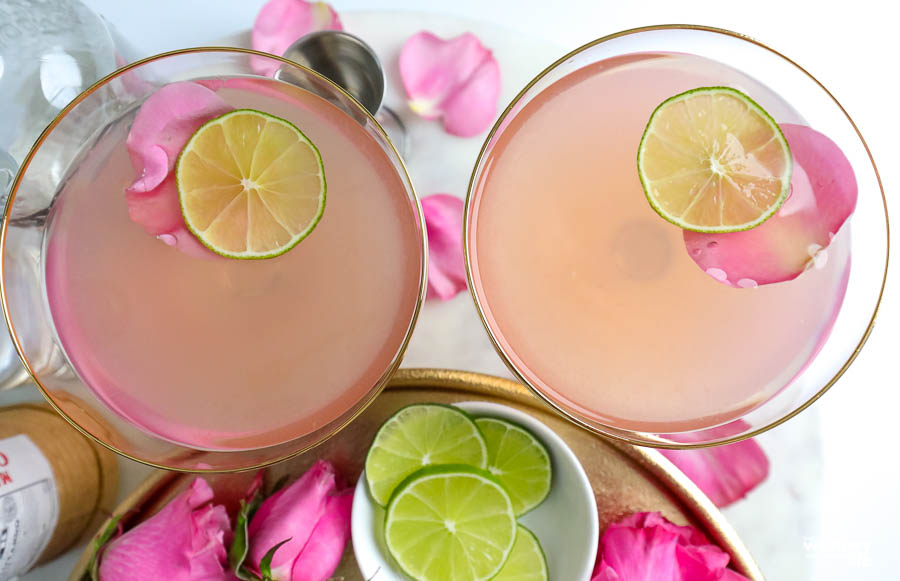 two cocktail glasses filled with a pink drink with roses petals and lime