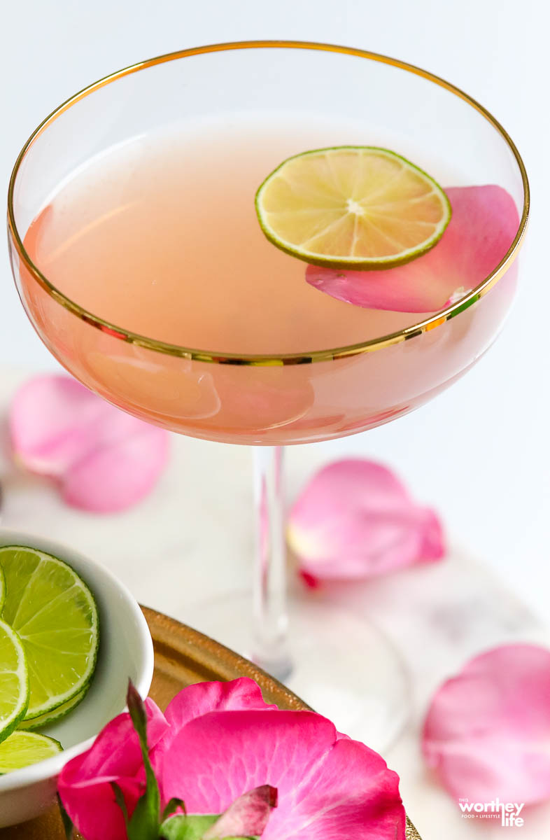 a pink gin cocktail in a couple glass garnished with a rose petal and lime