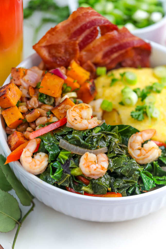 a white bowl filled with collard greens cheesy grits shrimp bacon and sweet potato hash