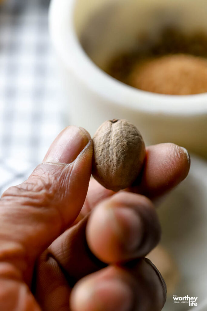 A hand holding a whole nutmeg in front of a small white bowl. 