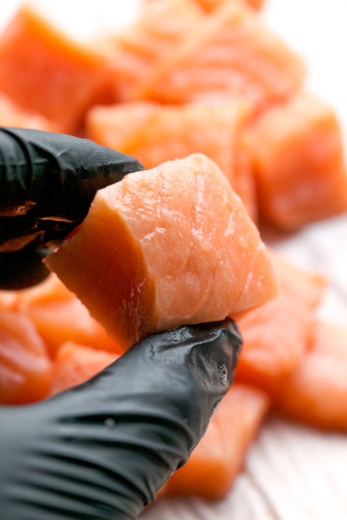 man with a black food glove on holding a fresh piece of cubed salmon
