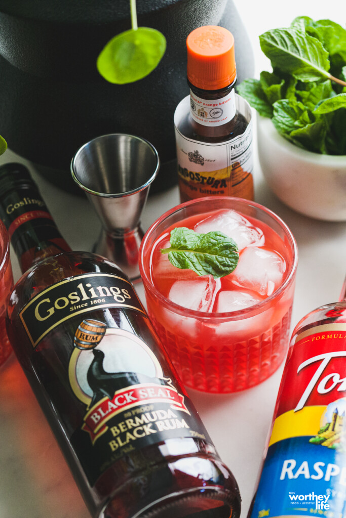 Raspberry Cocktails made with Rum next to a bottle of Bermuda Black Rum, a silver jigger and a bottle of bitters