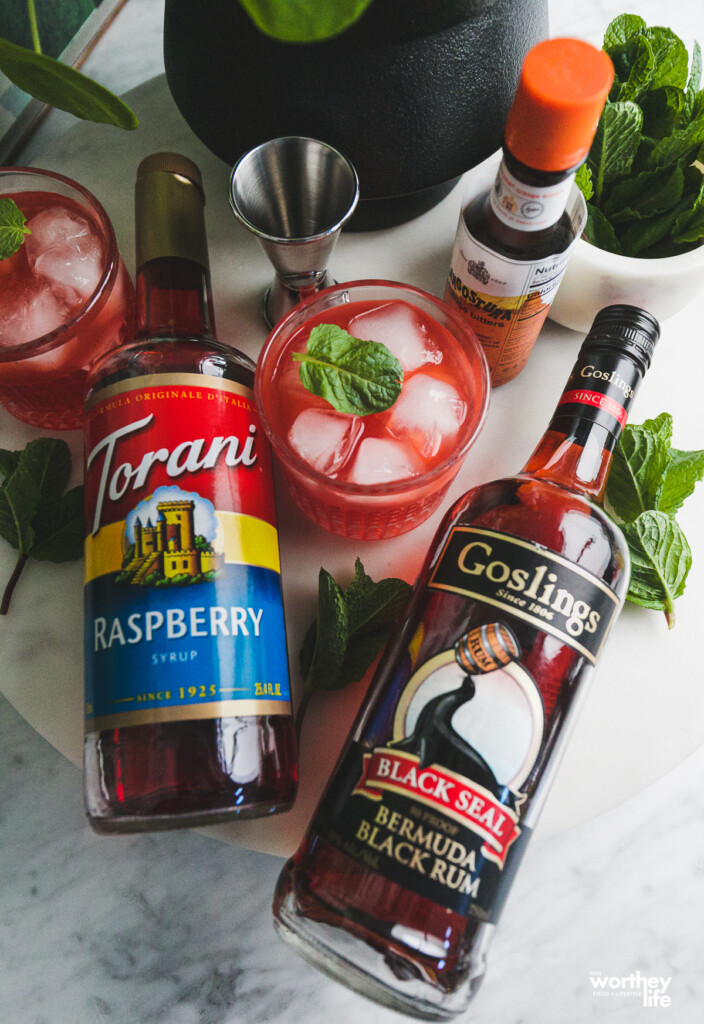 A bottle of Raspberry Torani, Goslings Black Seal Bermuda Black Rum, a raspberry cocktail, jigger and bitters shown on a white marble board