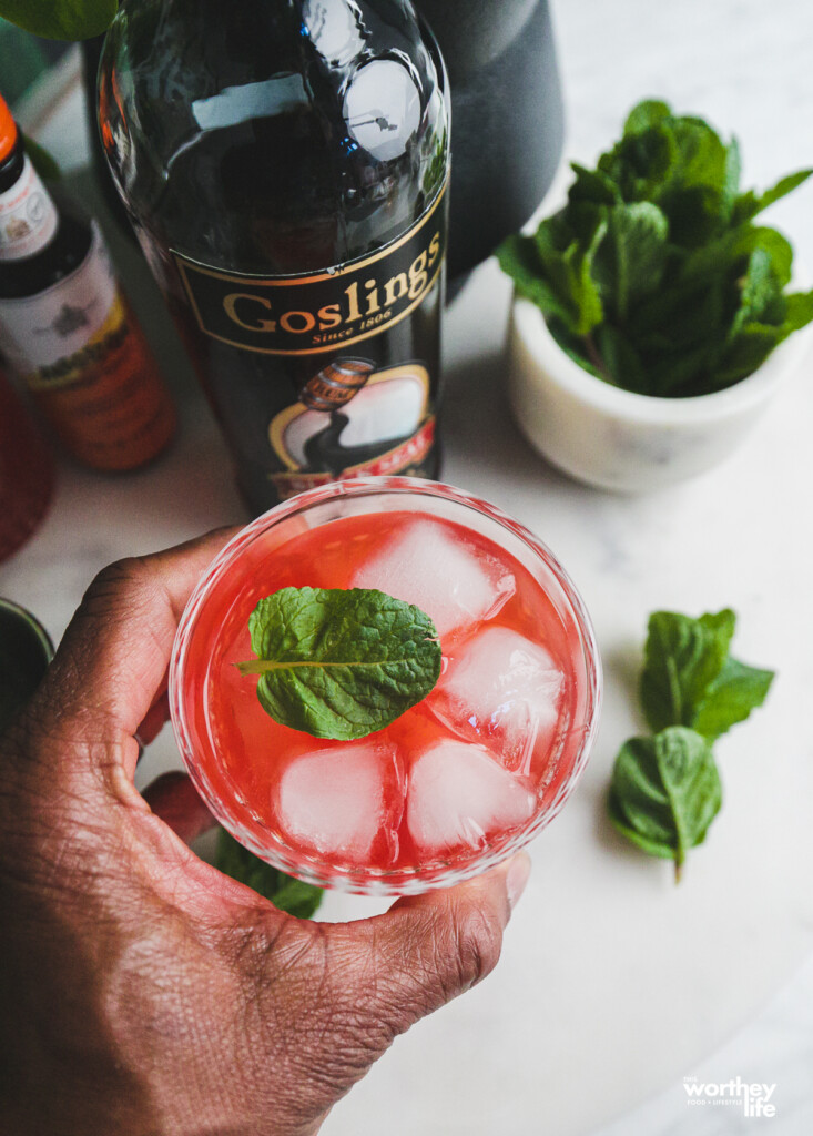 Man holding a Raspberry Cocktail in his hand. In the background is a bottle of Goslings Black Seal, next to a small white marble bowl of fresh mint. 