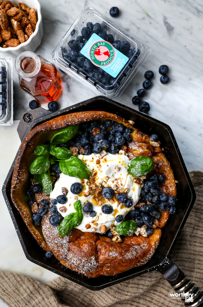 Blueberry Dutch Baby in a skillet with a pint of fresh blue berries, syrup, and praline pecans. 