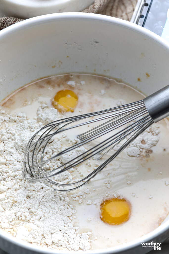 A large white bowl with Dutch Baby batter and a wire whisk. 