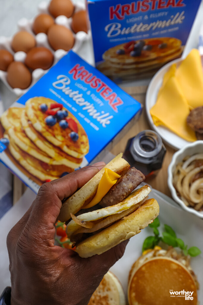 Holding a breakfast sandwich, with a box of pancake mix and toppings in the background. 
