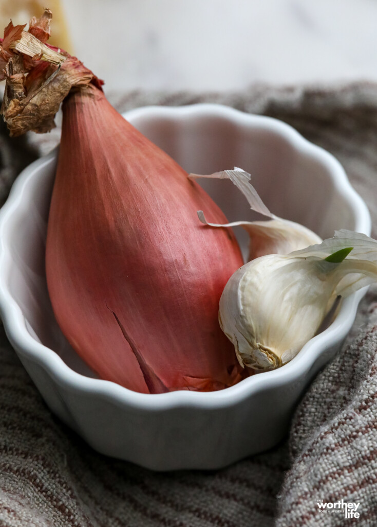A large shallot and two garlic cloves in a decorative white bowl ramekin.