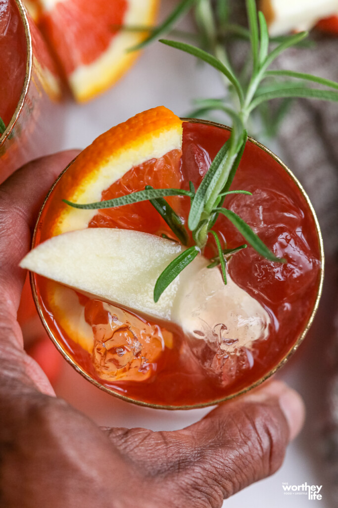A hand holding a cocktail garnished with apple, orange slices, and a sprig of fresh rosemary.