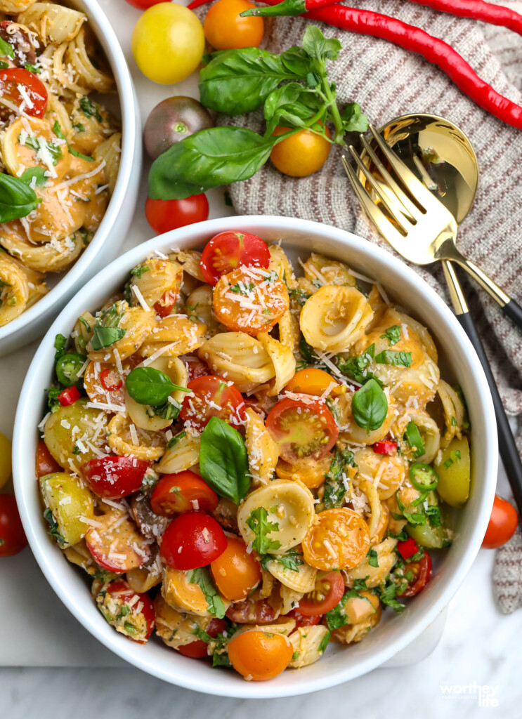 Two serving bowls filled with summer tomato pasta salad, fresh sweet basil on the side with loose tomatoes and a fork and spoon. 