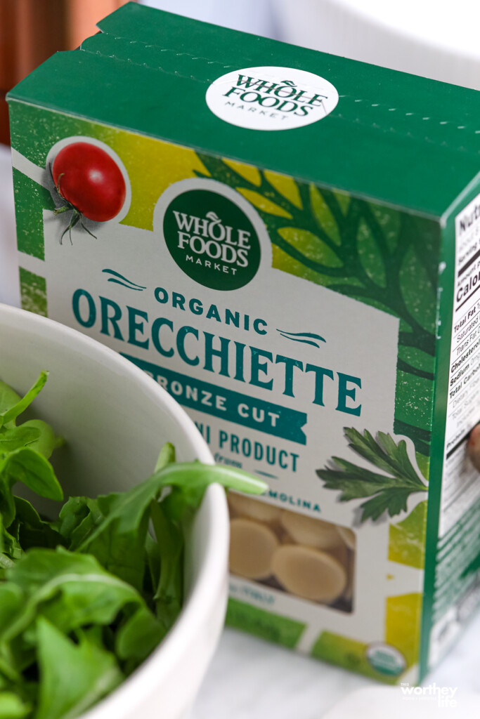 A box of orecchiette pasta with a white bowl filled with baby arugula in the foreground.