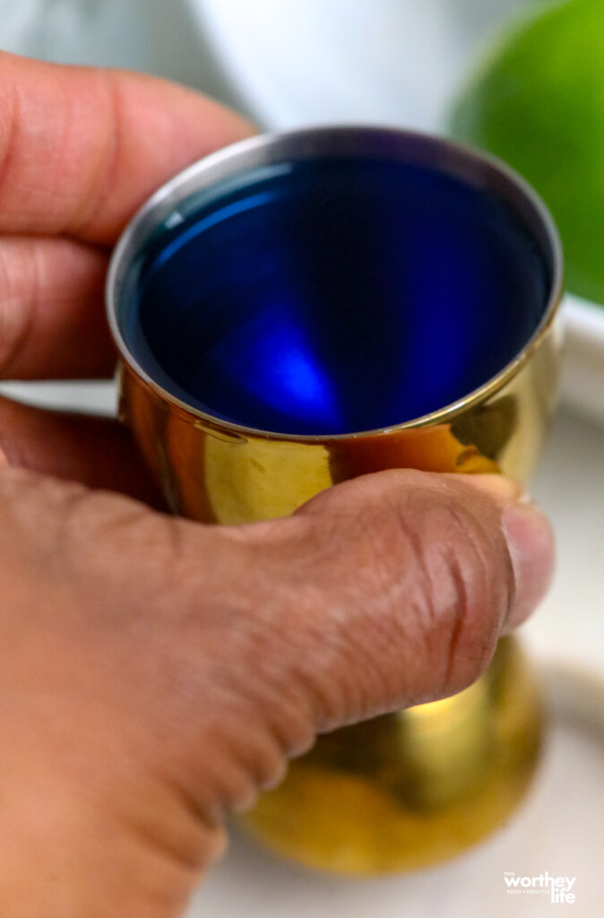 a man's hand holding blue syrup in a jigger