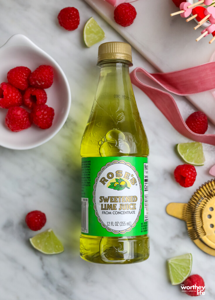 A bottle of sweetened lime juice with cut lime pieces and fresh raspberries. 