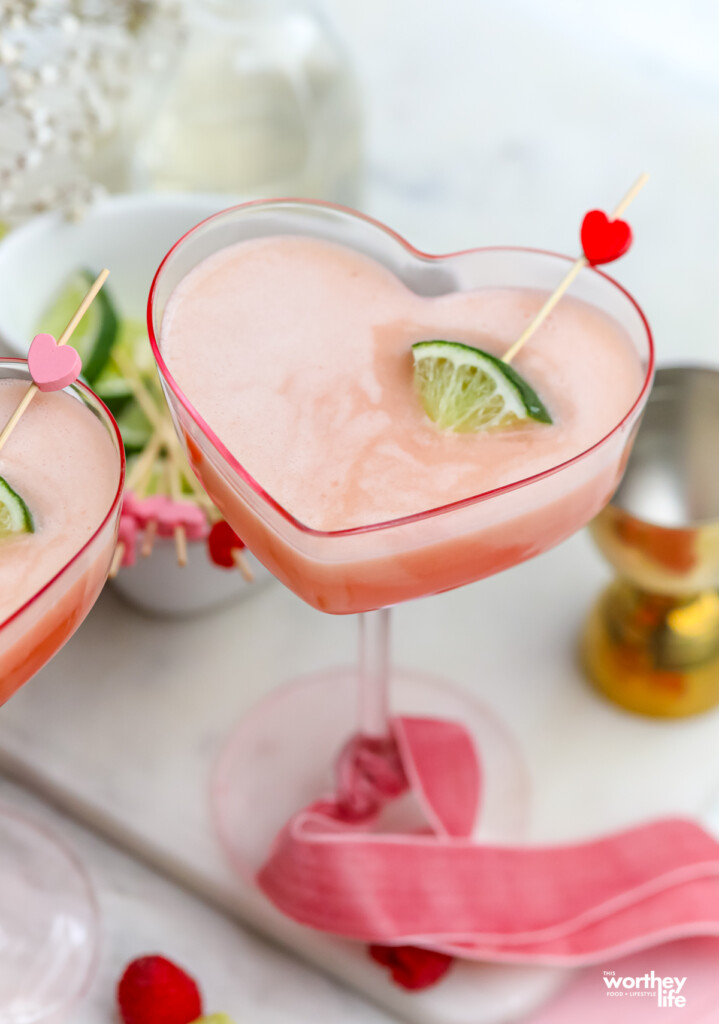 Our creamy raspberry cocktail on a white marble tray in a heart-shaped coupe with a pink ribbon. 