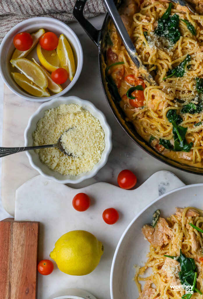 pasta in a pan with white bowls on the side filled with cheese and tomatoes and lemons