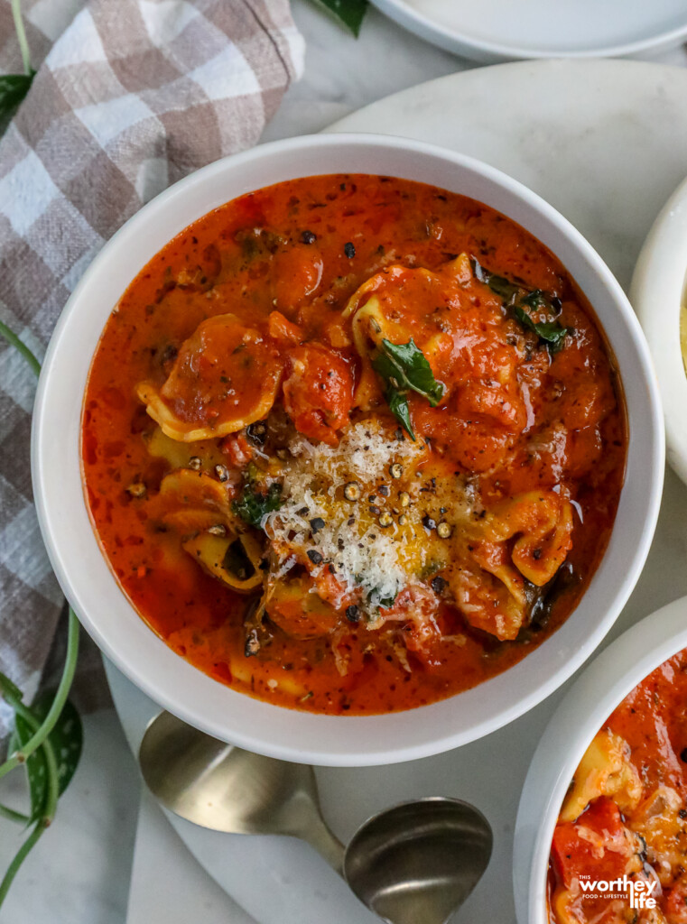 Tortellini Soup with Kale in a bowl with a white marble background, cloth napkin and gold spoons